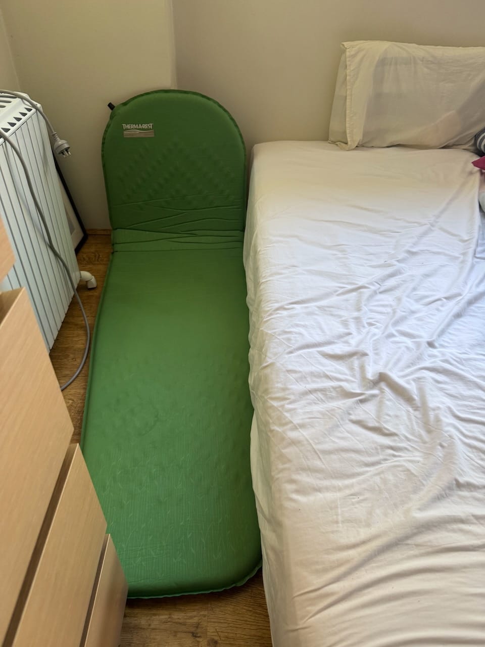 Green Thermarest brand camping mat, partially folded against the wall, flush against a bed side. A heater is visible on the left, and drawers are at the foot of the camping mattress.